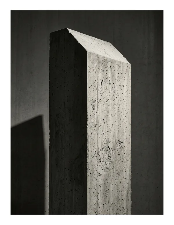 A tall monolithic concrete slab with a peaked top casts a dramatic shadow on a dark wall.